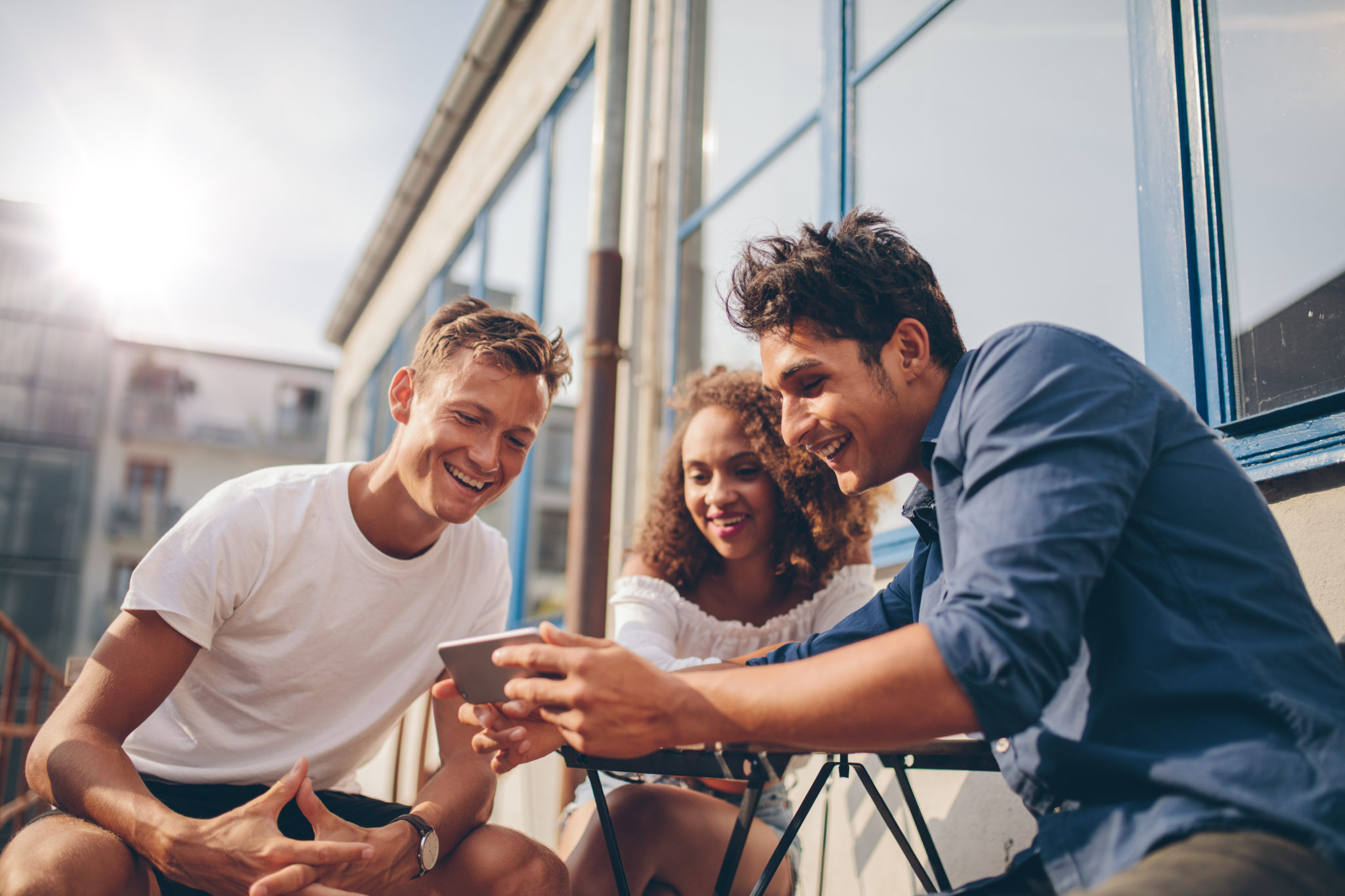 Três amigos novos que sentam-se ao ar livre e que olham o telefone móvel. Grupo de pessoas sentadas no café ao ar livre e assistindo vídeo no smartphone.